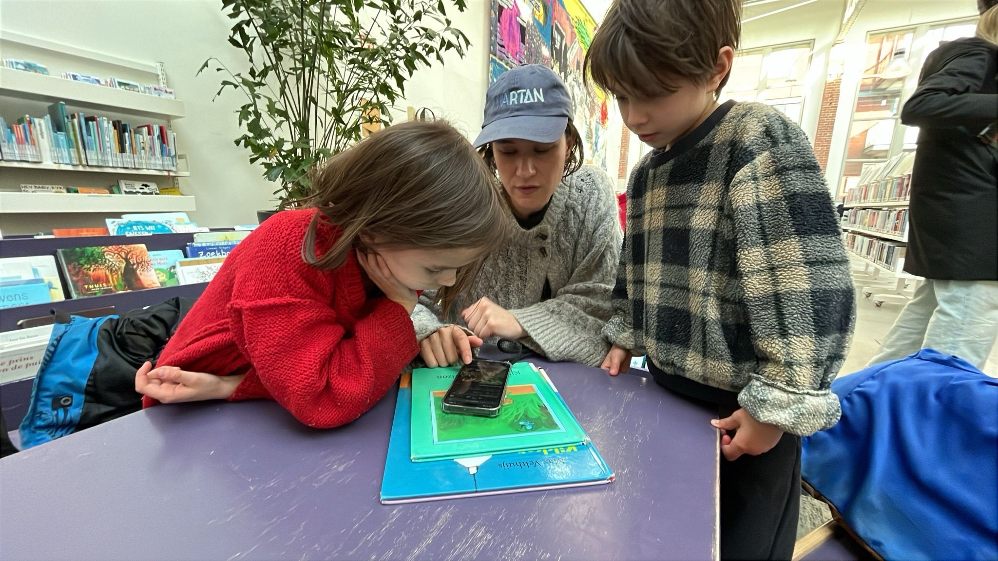 Een moeder en twee kinderen luisteren samen naar een telefoon aan een tafel in de bibliotheek